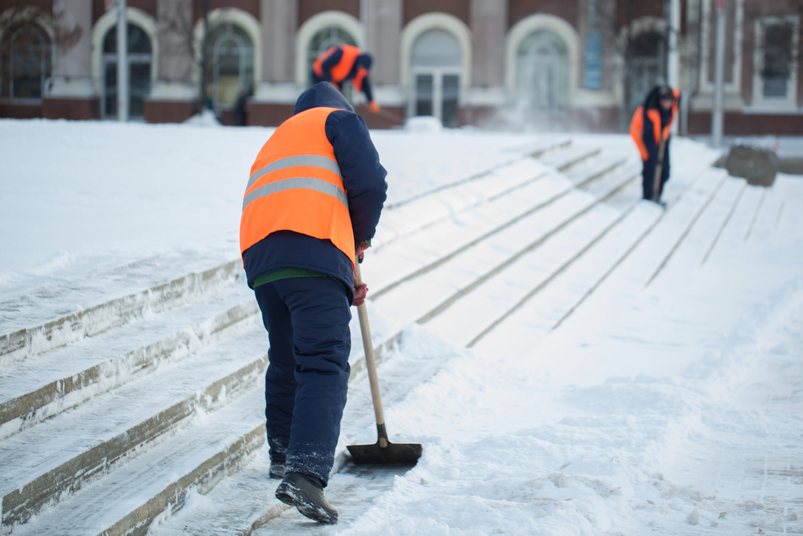 Workers shovel snow from steps and walkways outside a Denver building.